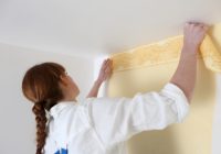 Frau mit braunen Haaren und Zopf bringt eine gelbliche Tapete an einer weißen Wand an.